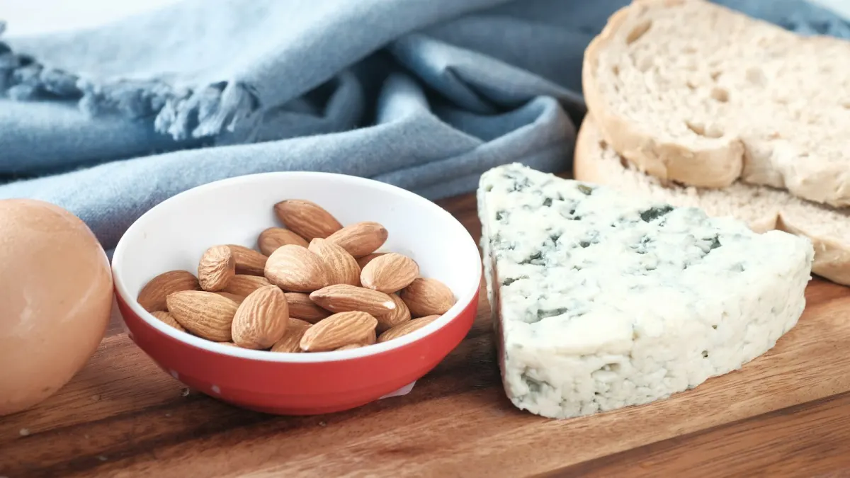 Een ei, een schaaltje amandelen, een stuk blauwe kaas en sneetjes brood op een houten snijplank