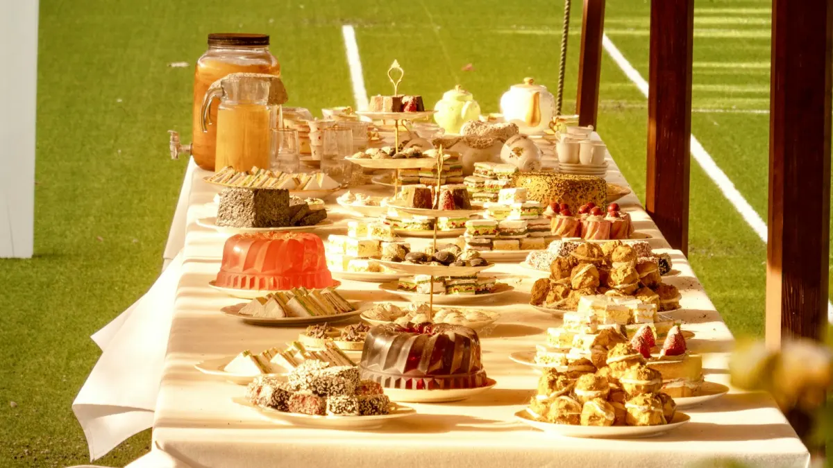 Lange tafel buiten in de zon vol taarten, cakes, soesjes en koekjes, met kannen limonade en theepotten