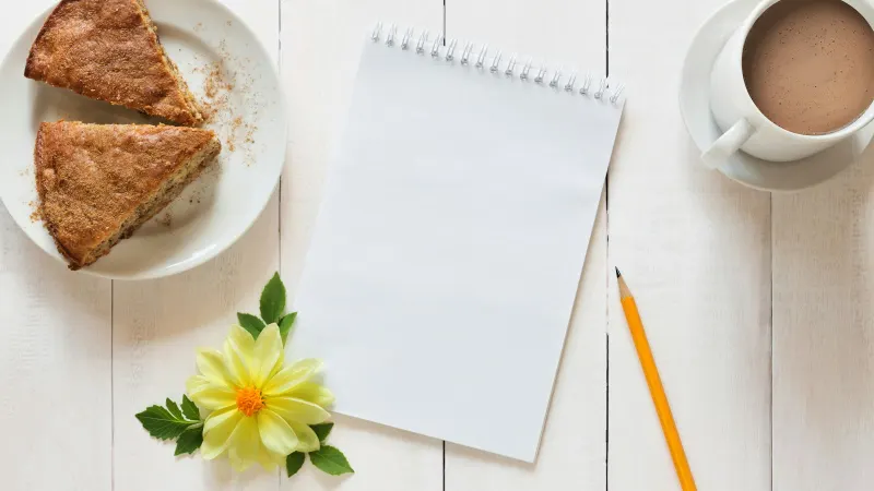 Twee stukken cake op een bordje naast een leeg notitieblok, een potlood, een kop koffie en een gele bloem