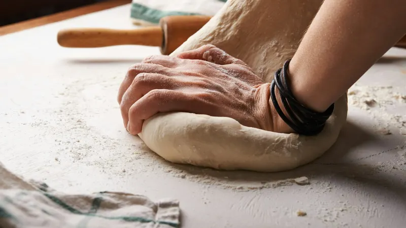 Handen kneden een bal brooddeeg op een met bloem bestoven werkblad, met een deegroller en een theedoek ernaast